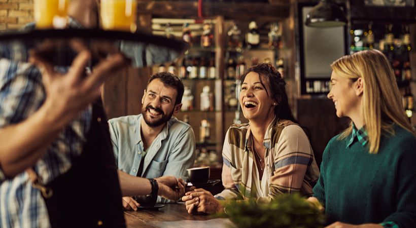 Cheerful friends having fun while talking to waiter in bar area.