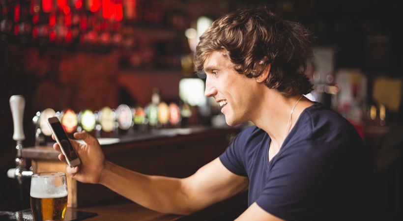 Man with beer glass using mobile phone at pub counter.