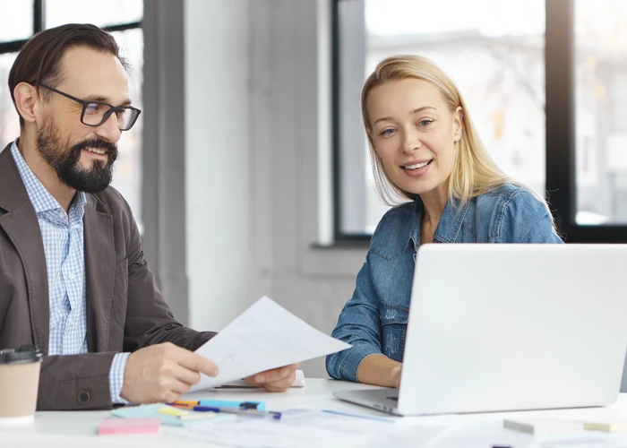 Blonde female manager and bearded male colleague reviewing documents together at a laptop in a bright office